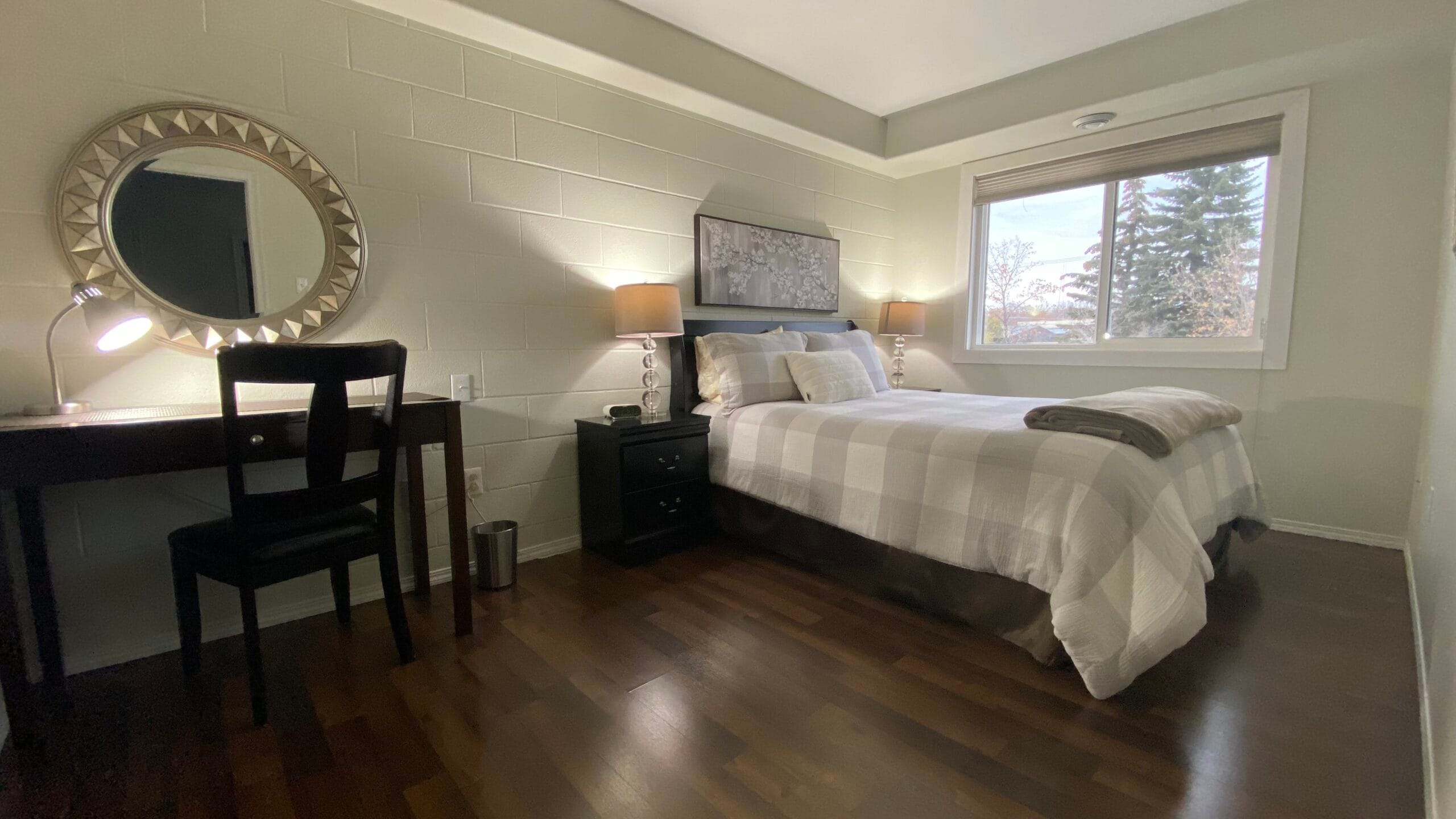 Queen bed with pretty bedding, two nightstands, two lamps, desk, desk lamp, chair, mirror, and window